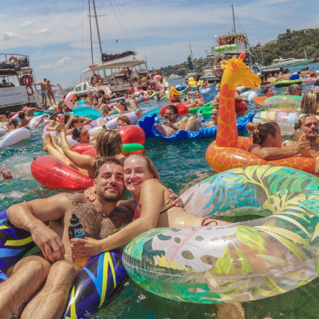 A crowd of people relaxing on colorful inflatable pool floats in the water at a lively boat party, with boats anchored nearby under a sunny sky. Two people smile at the camera in the foreground.