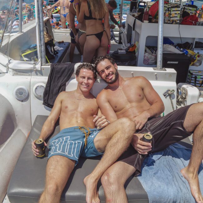 Two men are relaxing shirtless on a boat, smiling and holding drinks. There are other people in swimsuits and more boats on the water in the sunny background, suggesting a lively party atmosphere.