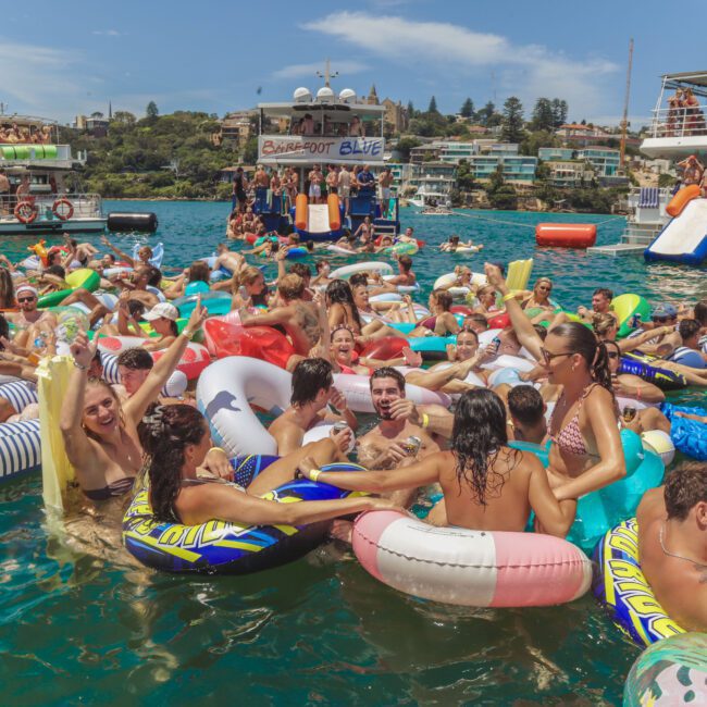 A lively crowd of people in swimsuits float on colorful inflatable tubes, partying and socializing in the water near boats on a sunny day. The scene is festive and crowded, with blue sky and greenery in the background.