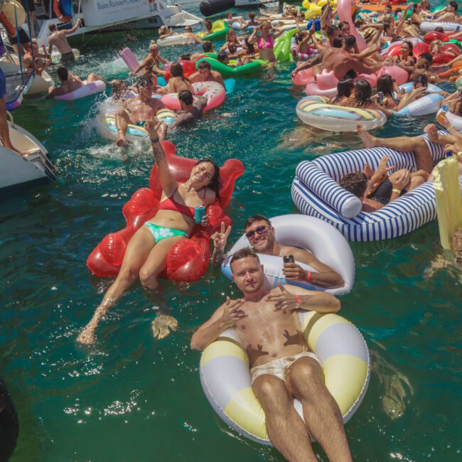 A large group of people relax and party on colorful pool floats in the water near boats, holding drinks and smiling under bright sunshine at a lively summer gathering.