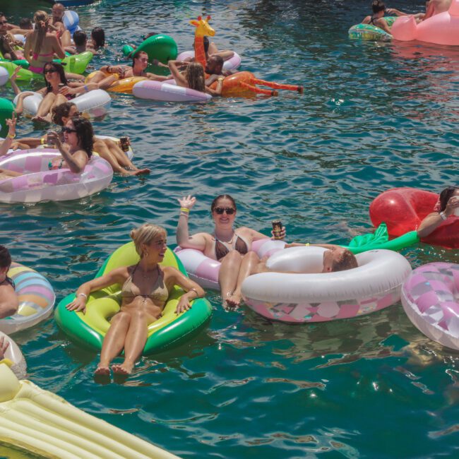 A group of people relax on colorful inflatable pool floats in a sunny outdoor setting, enjoying the water and socializing. Some hold drinks, and many are smiling and waving at the camera.