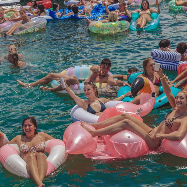 A large group of young people enjoy a pool party, lounging on colorful inflatable floats, including flamingos and unicorns, while others swim and smile in the water under bright sunlight.