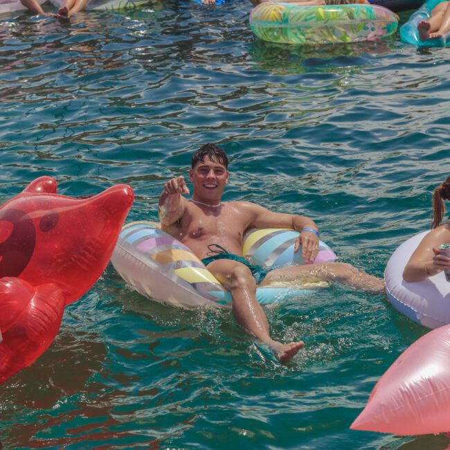 A group of young adults float on colorful inflatables in a sunny, open body of water. A smiling man in the center points toward the camera, surrounded by others relaxing and enjoying the event.