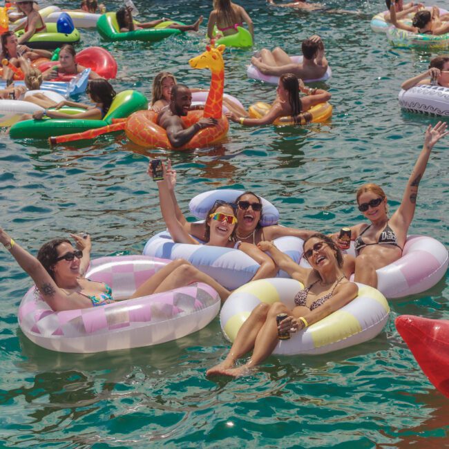 A group of people relax on colorful inflatables in a crowded pool, smiling and raising their arms in celebration. Others float nearby on various inflatable toys, enjoying the sunny day and festive atmosphere.