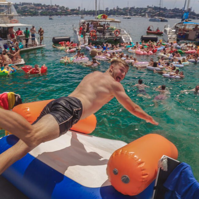 A man in swim trunks dives off a yacht onto an inflatable in the water, with many people on floats and boats celebrating together in a sunny harbor.
