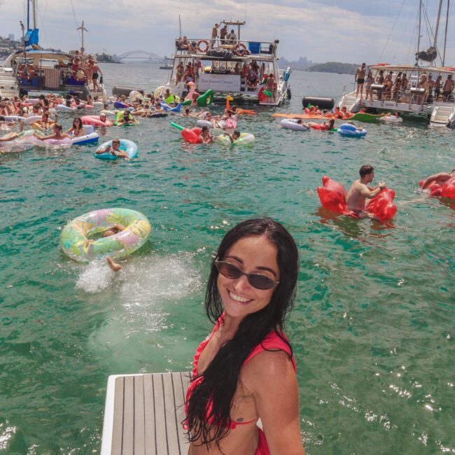 A woman in a pink bikini and sunglasses smiles while holding a drink on a boat deck. Behind her, people float and swim in a green-blue harbor, surrounded by boats under a cloudy sky.