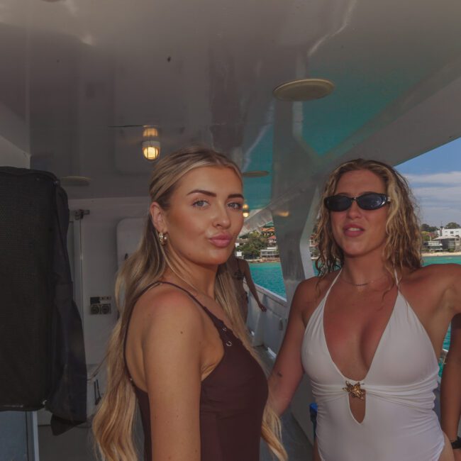 Three women enjoying a party on a boat, with one holding up a drink. The background shows blue water and houses along the shoreline. The mood is festive and relaxed.