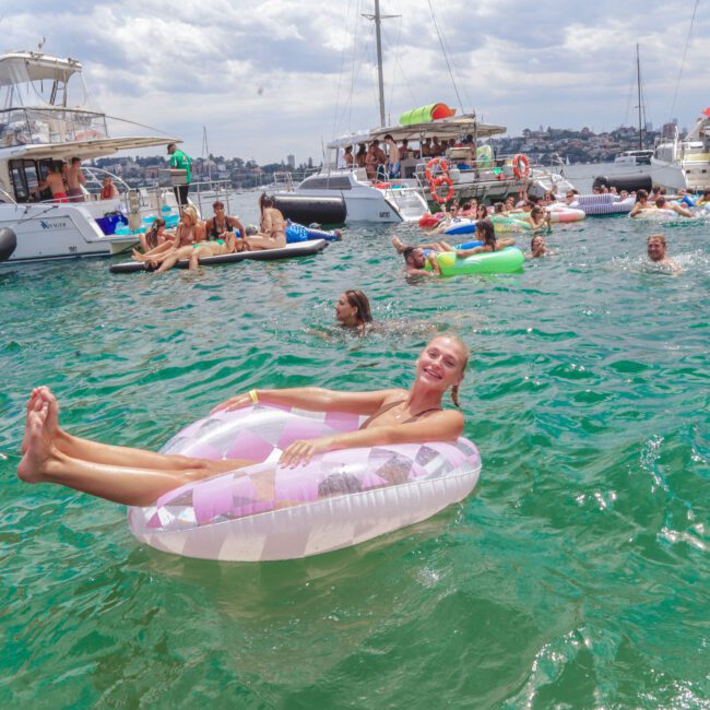 A woman smiles while reclining on a pink and white inflatable pool float in clear green water, surrounded by many people on floaties and boats under a partly cloudy sky.