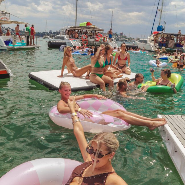 A group of people relax on inflatable floats and swim in a marina, surrounded by yachts and boats under a partly cloudy sky. Some lounge on a dock while others enjoy the water, creating a lively summer scene.