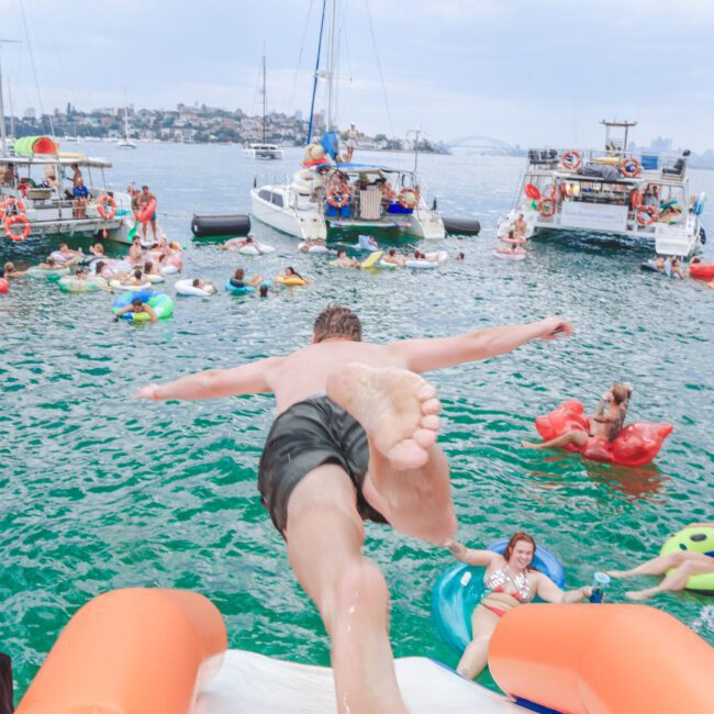 A man in swim trunks jumps off a boat into the water, surrounded by people relaxing on colorful inflatables and nearby boats on a lively day out on the harbor.