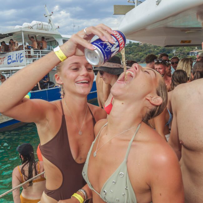 A woman in a brown swimsuit pours a drink from a can into another woman’s mouth at a crowded boat party. People around them are smiling and enjoying the lively scene on the water.