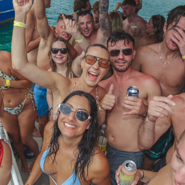 A group of young adults in swimsuits smile, laugh, and pose with drinks on a crowded boat, enjoying a lively party on the water. The atmosphere is festive and energetic.
