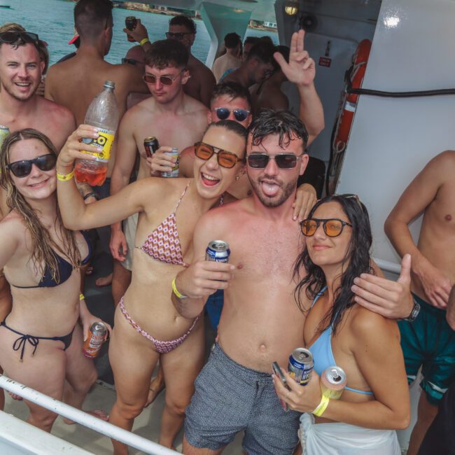 A group of young adults in swimwear pose and smile on a yacht, holding drinks and enjoying a sunny day on the water. Some make hand signs and wear sunglasses. The atmosphere is festive and carefree.