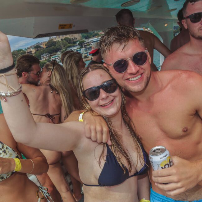 A group of young adults in swimsuits enjoy a lively boat party. Two people in the center smile at the camera, arms around each other, holding drinks. Others stand nearby, and the ocean and shoreline are visible in the background.