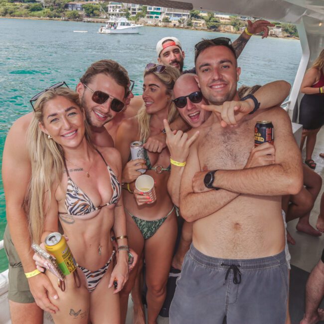 A group of young adults in swimsuits smile and pose together on a boat, holding drinks. The boat is docked near clear turquoise water with more boats and a shoreline visible in the background.