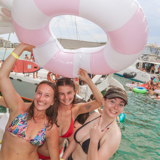 Three smiling women in swimsuits pose on a boat holding a large pink and white inflatable ring, with other people on boats and inflatables enjoying the water in the background on a sunny day.