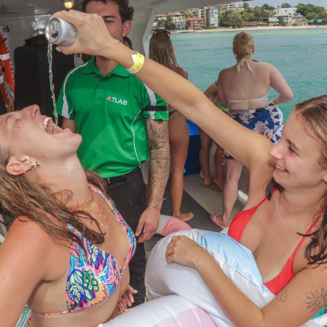 Two women in swimsuits on a boat; one woman is pouring a drink from a can into the other's mouth as they laugh. People in swimwear and the ocean are visible in the background.