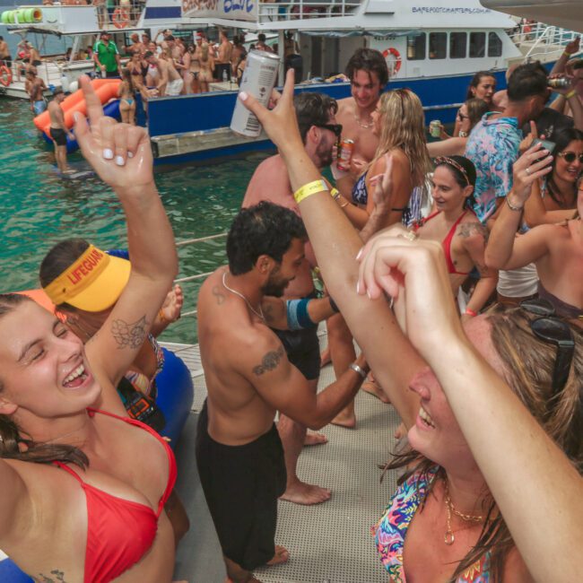 A group of young adults in swimwear dance, laugh, and celebrate on a boat. The party is lively, with people holding drinks and raising their hands. Other boats and people are visible in the water nearby.