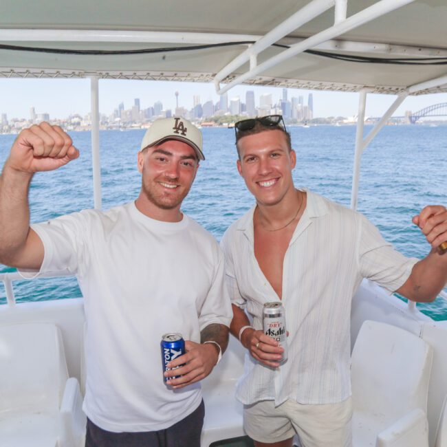 Two men smiling and holding drinks stand on a boat with a city skyline and a bridge in the background. Both are wearing casual summer clothes and posing with one arm raised in celebration.