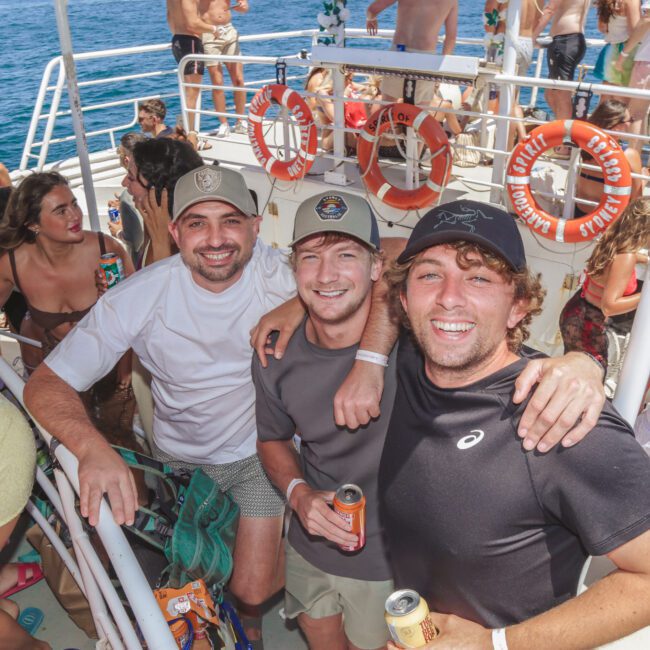 Three men smiling and posing together on a crowded boat party, surrounded by other people enjoying themselves. The boat is on the water under sunny skies, with life rings and railings visible.