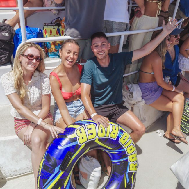 Three smiling people sit on stairs outdoors, holding a blue inflatable tube labeled “Rider.” They wear summer clothes and sunglasses, and other people with bags and coolers stand nearby, suggesting a lively, sunny gathering.