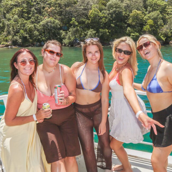 Five women in swimsuits and summer clothes stand smiling on a boat in sunny weather, holding drinks, with green water and trees in the background.