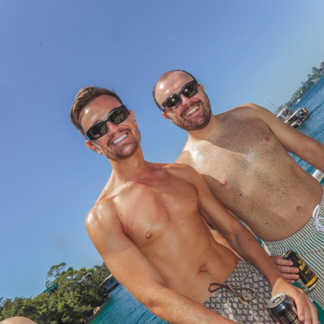 Two men in swim trunks and sunglasses smile at the camera while standing on a boat. They are holding cans and enjoying a sunny day by the water, with trees and boats visible in the background.