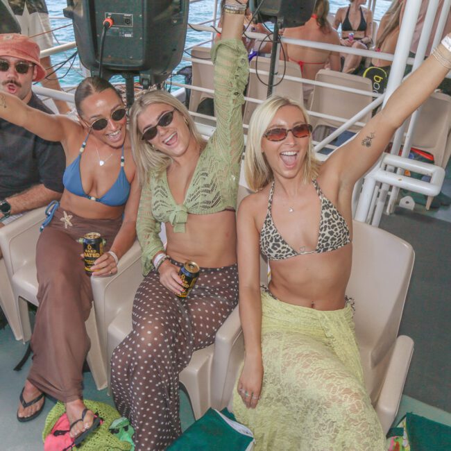 Three women in swimsuits and sunglasses sit together smiling and raising their arms, holding drinks, at a lively boat party, with other people and water visible in the background.