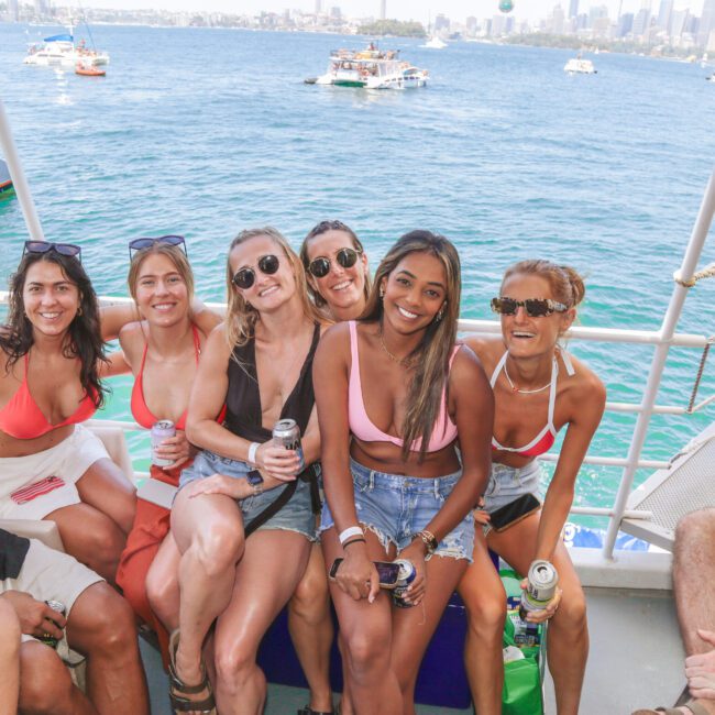 Six women in swimsuits and sunglasses smile and pose together on a boat with drinks in hand. The background shows blue water, other boats, and a distant city skyline on a sunny day.
