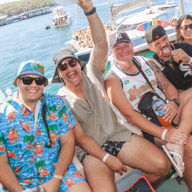 A group of five smiling friends in casual summer clothes sit on a boat, enjoying a sunny day on the water with blue seas and other boats in the background. One person raises a hand in excitement.