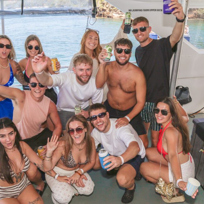 A group of twelve young adults pose and smile on a boat, holding drinks and wearing swimwear. The sun is shining, and water and a rocky shoreline are visible in the background. Everyone appears happy and energetic.