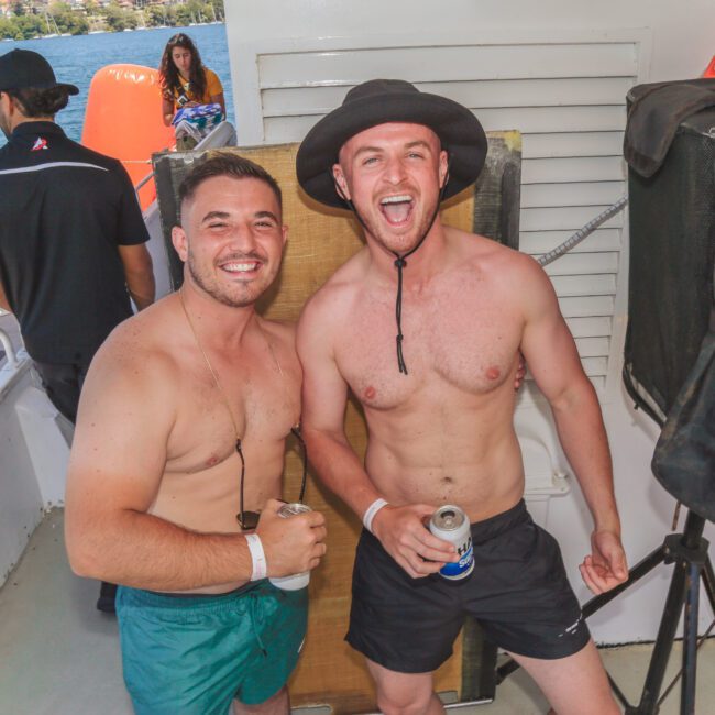 Two smiling men in swim trunks stand on a boat holding drinks. One wears a sunhat and both have wristbands. Other people and water are visible in the background; it’s a sunny day.