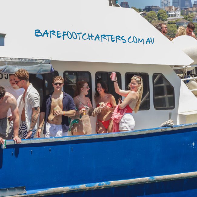A group of young people in swimwear stand and socialize on the deck of a blue and white charter boat called Barefoot Charters, with others relaxing in the background on a sunny day.