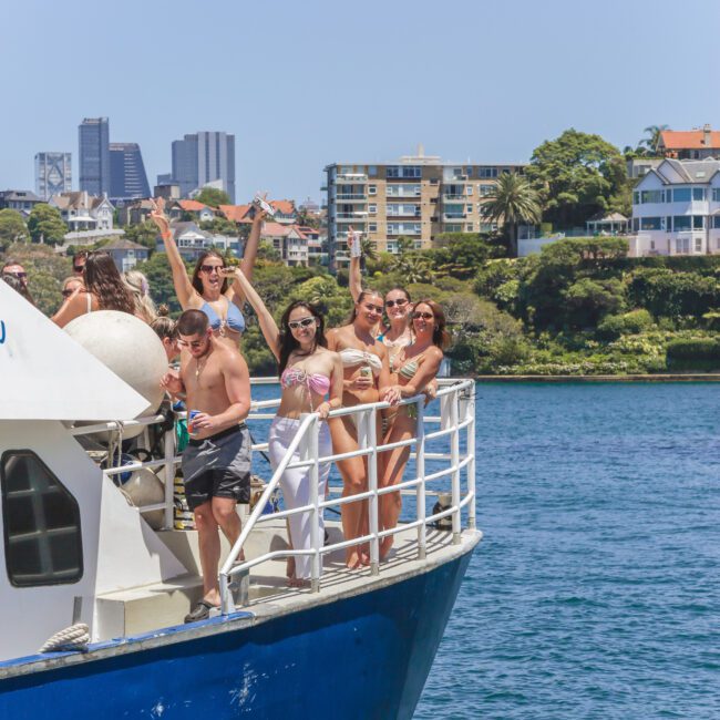 A group of people in swimwear stand and pose cheerfully at the bow of a blue and white boat on the water, with city buildings and greenery visible in the background on a sunny day.
