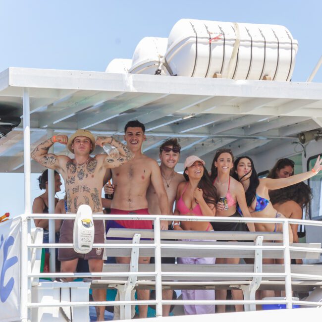 A group of young adults in swimwear smile and pose for a photo on the deck of a boat during a sunny day. Some flex their arms and others lean together, enjoying a fun outing. A stuffed giraffe toy is visible in the background.