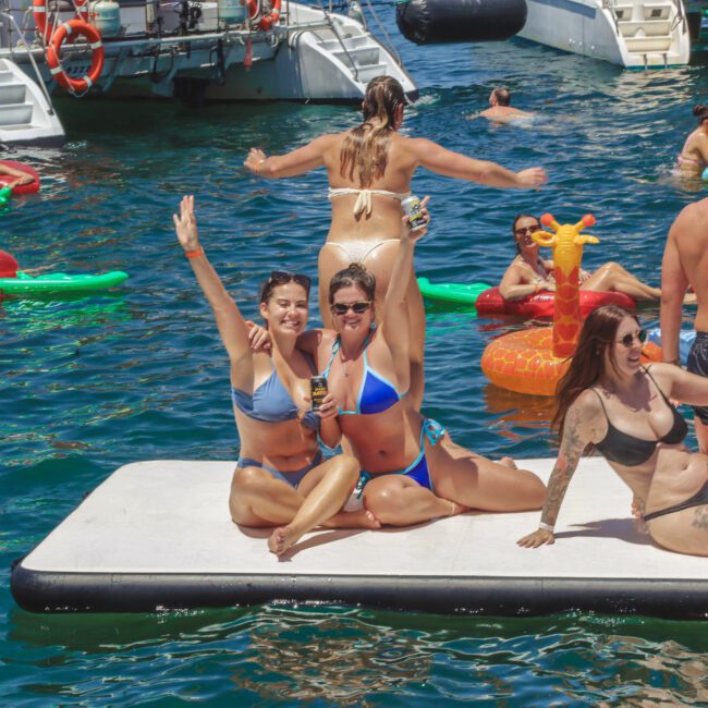 People in swimsuits enjoy a lively pool party on a floating platform and inflatables in the water, with boats in the background. Two women in blue bikinis sit in front, smiling and waving at the camera.
