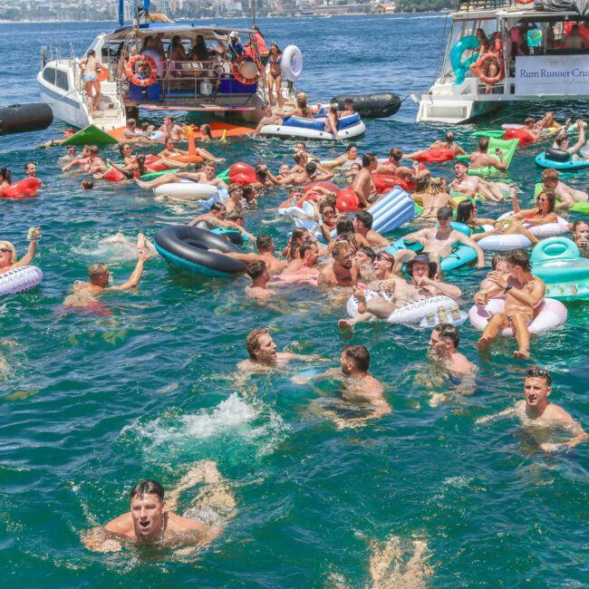 A large group of people swim and float on colorful inflatables in the ocean near several anchored boats during a lively summer party, with clear skies and water in the background.