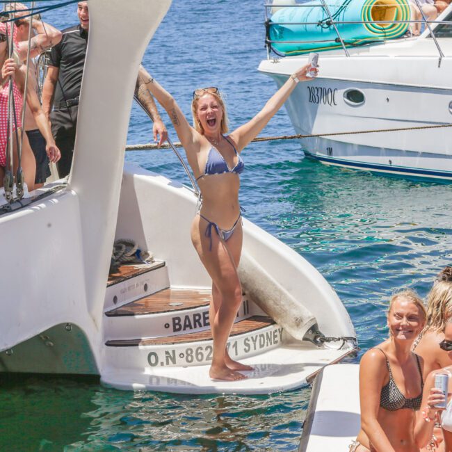 A woman in a bikini stands on the back of a boat, arms raised and smiling, surrounded by friends. Other people relax nearby, and another boat floats in the background on a sunny day.