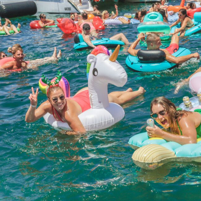 A group of people relax on colorful floaties, including a unicorn, in bright blue water. They are smiling, wearing swimsuits, and enjoying drinks at a lively outdoor pool or lake party.