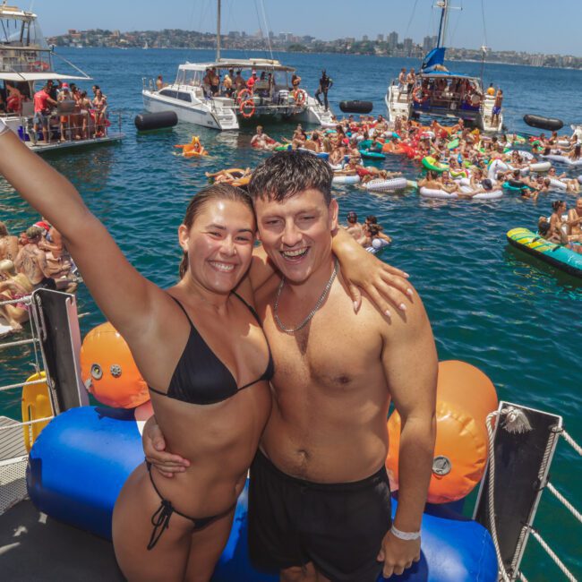 A smiling woman and man in swimsuits pose on a boat, with the woman raising one arm. Behind them, many people float on inflatables and swim near several boats on a sunny day.