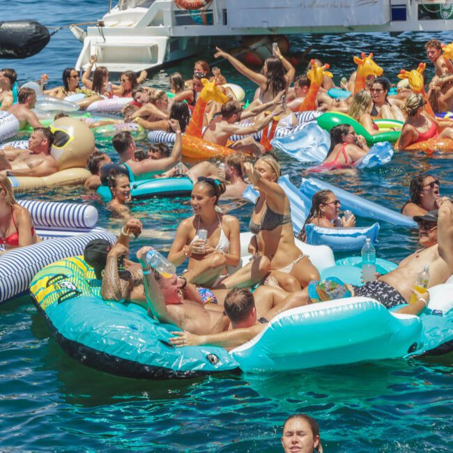 A large group of people relaxing on colorful inflatable floats and swimming in clear blue water during a lively pool party near a boat under sunny weather.