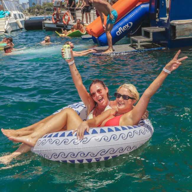 Two women in swimsuits smile and pose with raised arms while sitting together in a patterned inflatable tube on clear blue water at a lively pool or yacht party. One holds a drink, and people are swimming and playing in the background.