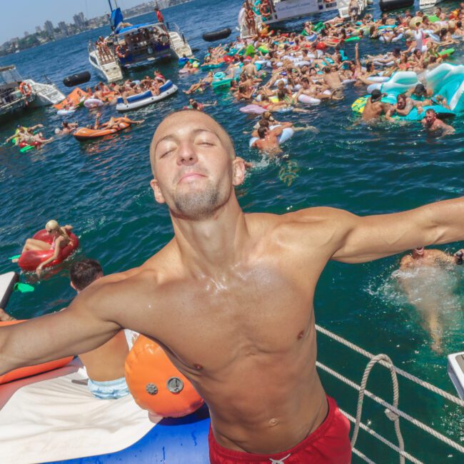 A man with arms outstretched stands at the edge of a boat, eyes closed, surrounded by people swimming and relaxing on inflatables in the water, with other boats and a city skyline in the background on a sunny day.