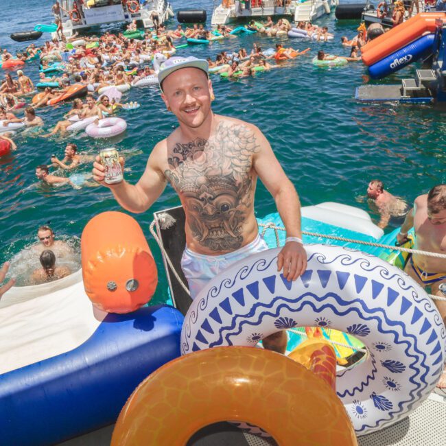 A smiling man with tattoos, holding a drink, stands on a boat among colorful inflatable pool floats, with many people swimming and relaxing in the water at a lively summer party.