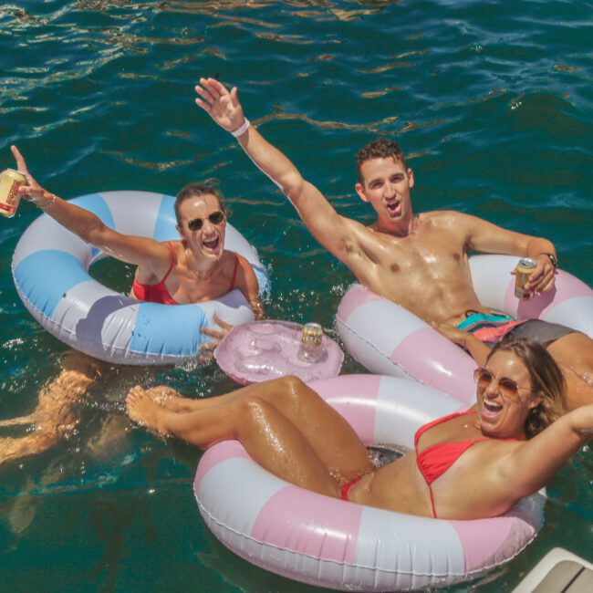 Three people in swimsuits float on colorful inflatables in blue water, smiling and raising their arms while holding drinks. The scene is sunny and cheerful, with a Yacht Social Club logo in the corner.