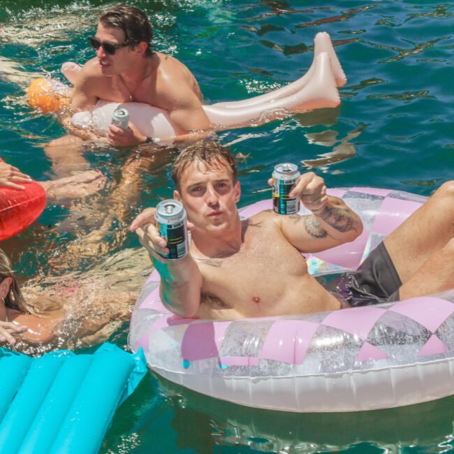 Four people relax on colorful pool floats in the water. A man in front holds up two cans and smiles at the camera, while others lounge nearby, enjoying drinks and the sunny weather. The "Yacht Social Club" logo is visible.