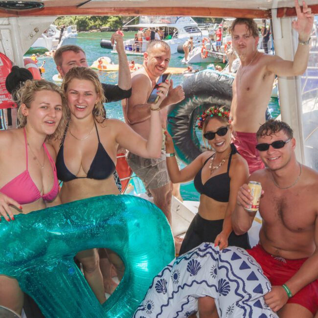 A group of young adults in swimsuits smile and pose on a boat, holding drinks and a large inflatable letter "D" with other boats and people in the water visible in the background. It’s sunny and festive.