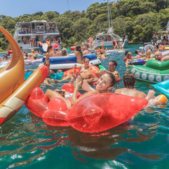 A large group of people float on colorful inflatable pool toys in blue water near boats, enjoying a sunny day with smiles and laughter. Green trees line the background shore.