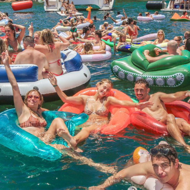 A large group of people relax and have fun on colorful inflatable floaties in a crowded, sunny lake scene. Everyone is in swimsuits, enjoying the water and lively atmosphere on a bright summer day.