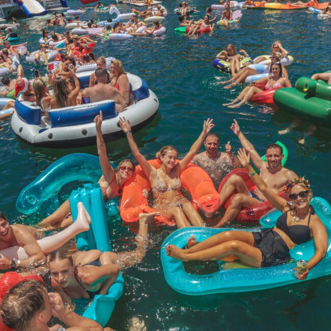 A large group of people relaxing on colorful inflatable floats in the water, smiling, waving, and enjoying a sunny day at a lively pool or boat party.